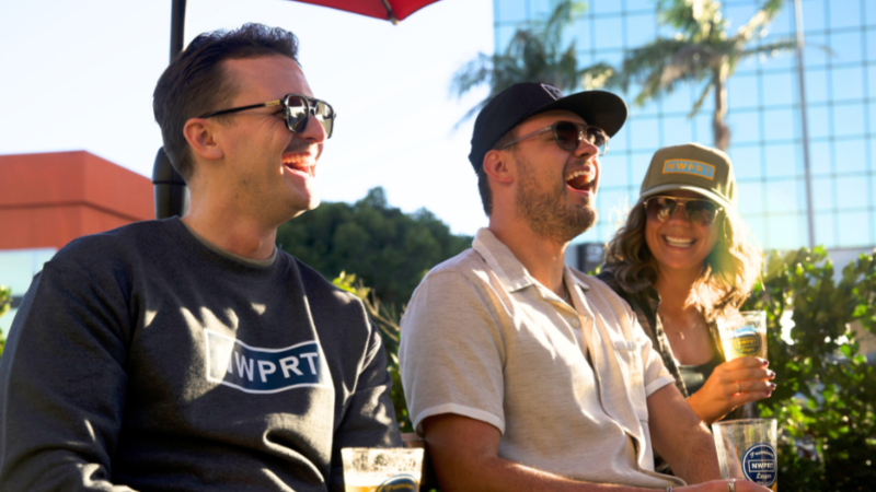 Three people sitting outdoors, enjoying NWPRT lager under an umbrella.
